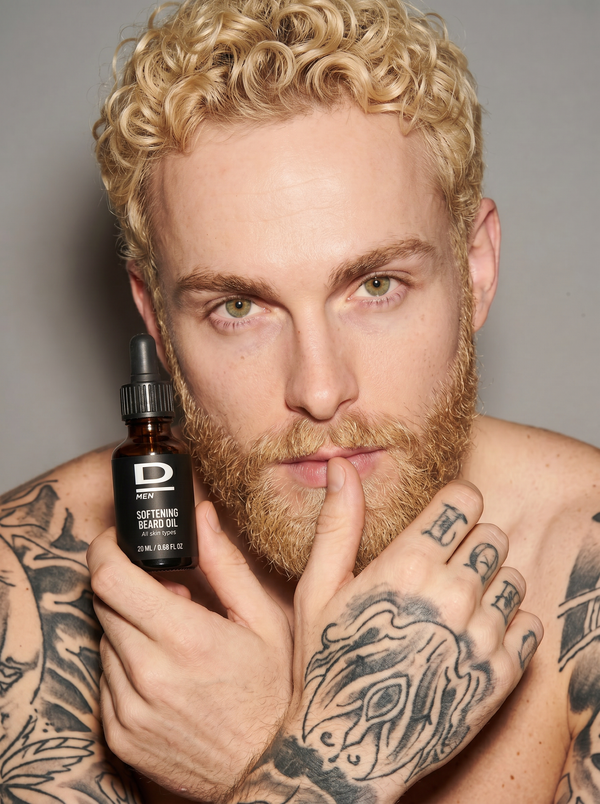 Bartträger mit blonden Locken und Tätowierungen hält eine Flasche mit der Aufschrift 'D MEN SOFTENING BEARD OIL' nah an sein Gesicht