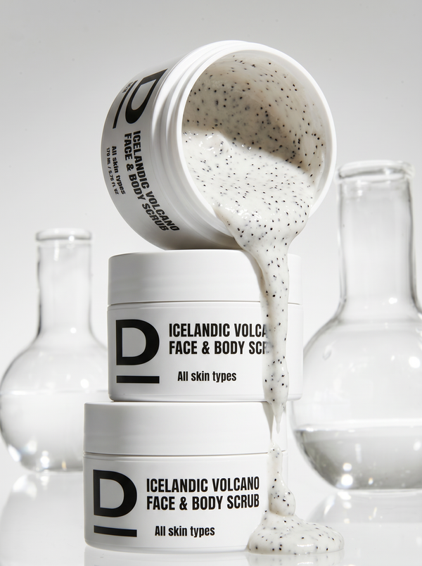 Drei weiße Tiegel mit schwarzer Aufschrift 'ICELANDIC VOLCANO FACE & BODY SCRUB All skin types', wobei aus dem oberen Tiegel eine cremige, körnige Substanz fließt, im Hintergrund zwei Glasgefäße mit klarem Inhalt