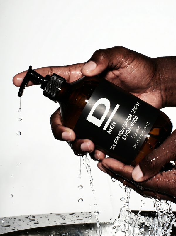 Hände halten eine braune Flasche mit Pumpspender, aus der Flüssigkeit tropft, auf der Flasche steht 'D MEN SILK SKIN BODY SERUM SPRAY SANDALWOOD DRY OIL 16 FL OZ'