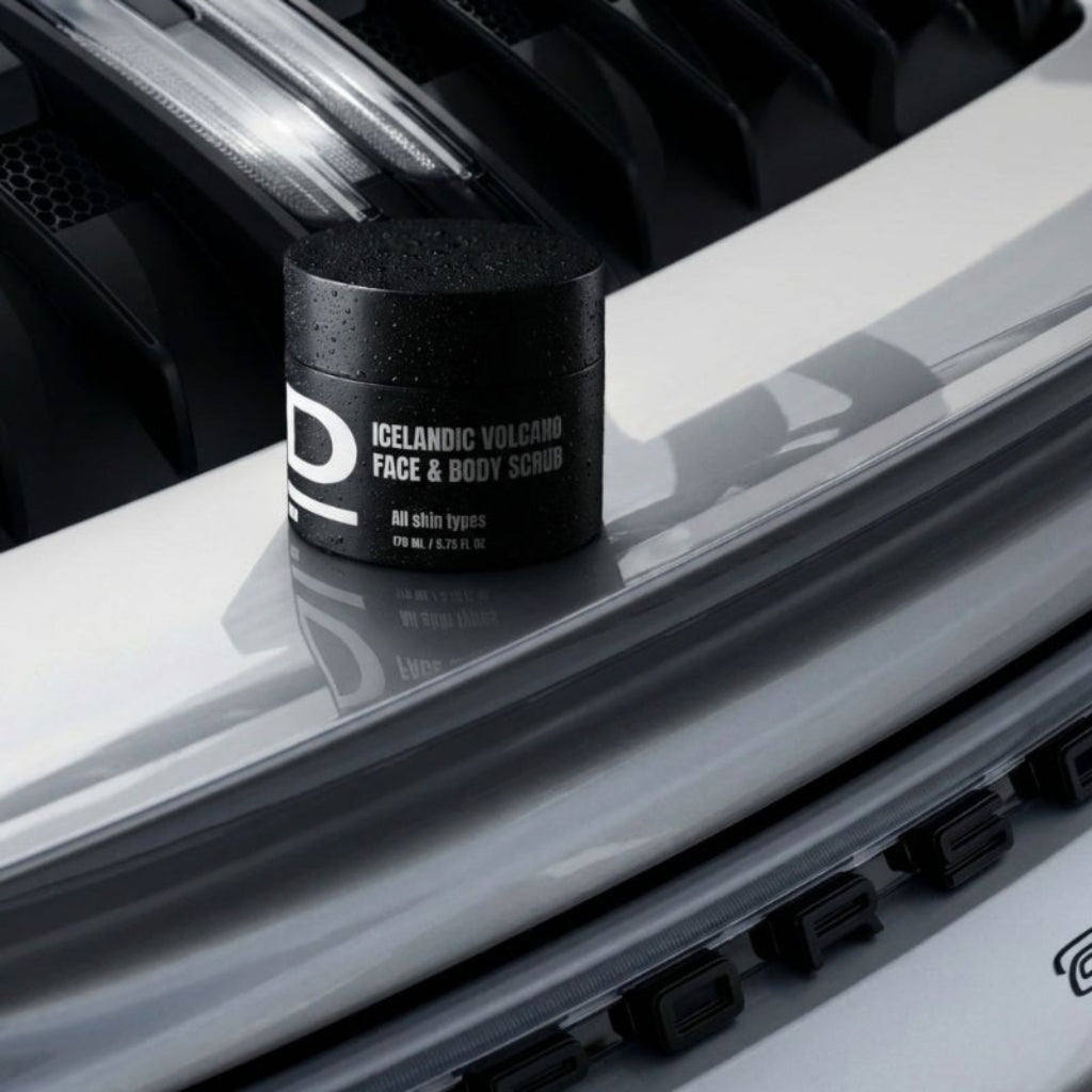 Schwarzer Tiegel mit der Aufschrift 'ICELANDIC VOLCANO FACE & BODY SCRUB' steht auf glänzend weißer Oberfläche mit Wassertropfen.