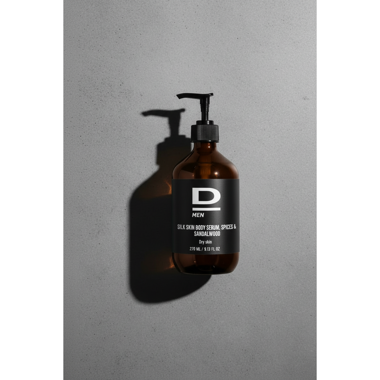 Braune Glasflasche mit Pumpspender auf grauer Oberfläche. Das schwarze Etikett trägt ein großes weißes D und darunter die Aufschrift 'MEN SILK SKIN BODY SERUM, SPICES & SANDALWOOD by dAlba 270 ml / 9.13 FL.OZ.'. Die Flasche wirft einen deutlichen Schatten nach links unten.