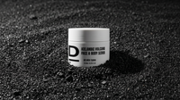 Weißer Tiegel mit schwarzer Aufschrift 'ICELANDIC VOLCANO FACE & BODY SCRUB' steht auf schwarzem, körnigem Vulkanboden