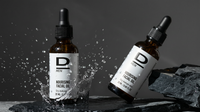 Zwei braune Glasflaschen mit Pipetten, beschriftet mit 'D MEN NOURISHING FACIAL OIL', stehen auf dunklem Stein, eine Flasche steht in einer Wasserspritzung