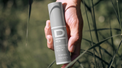 Hand hält zylindrischen Sonnenschutz-Stick mit Aufschrift 'D MEN SUN PROTECTION SPF50 STICK WITH TINT' vor unscharfem Hintergrund mit Gräsern